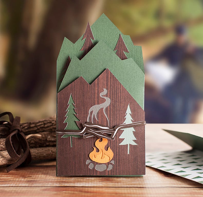 A handmade card decorated with trees and a campfire sits on a desk.