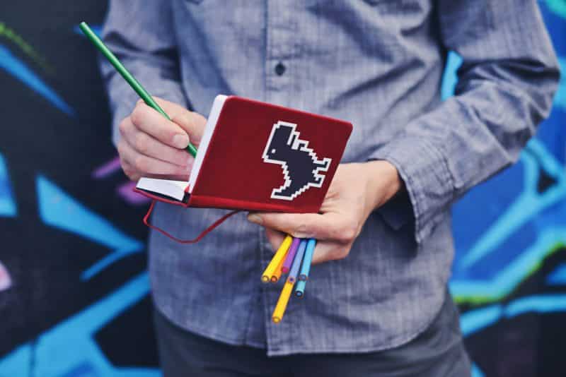 A man writes in a small red notebook decorated with a pixel dinosaur sticker