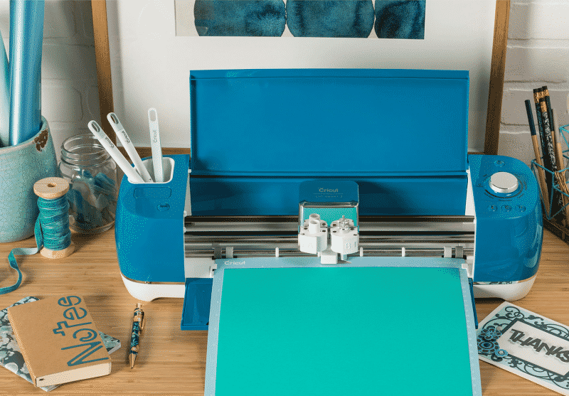 A bright Blue Cricut Explore Air 2 cuts vinyl on a desk full of crafting supplies.