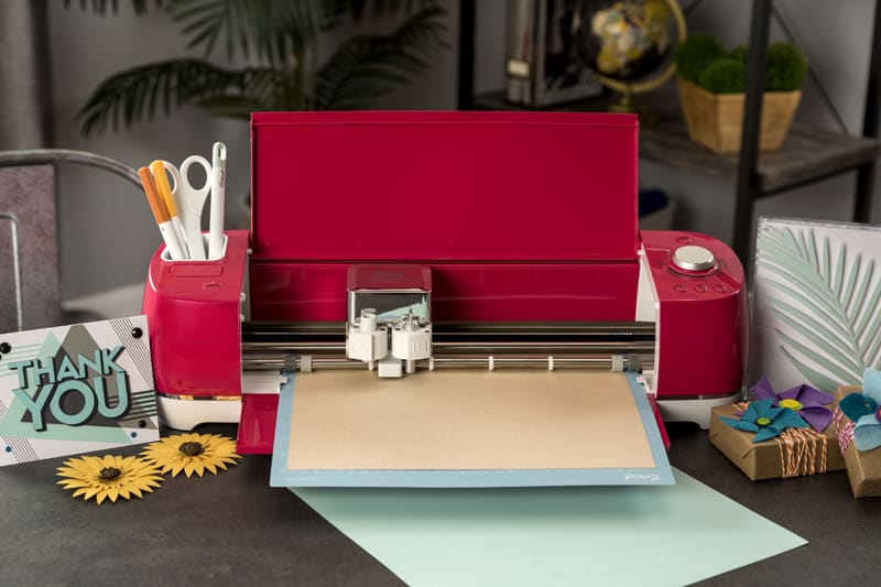 A raspberry Cricut Explore Air 2 sits on a desk cutting vinyl.